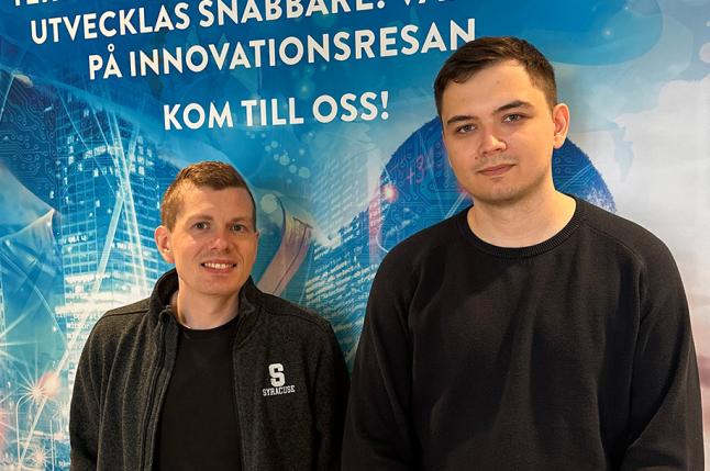 Seamus och Alexander, två nya medarbetare hos Infotiv står framför en blå reklamaffisch.