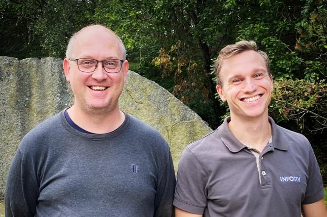 Två män står framför en stor sten och en grön skog. 