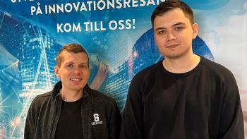 Seamus och Alexander, två nya medarbetare hos Infotiv står framför en blå reklamaffisch.